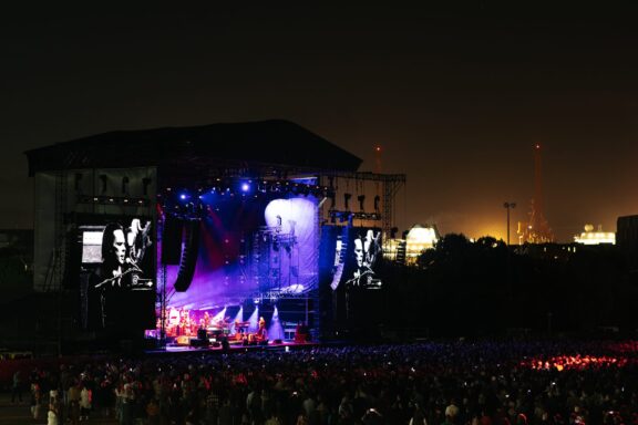 Large outdoor concert at night with a brightly lit stage, colorful lights, and video screens. A large crowd is gathered in front of the stage, with city lights and dark skies in the background.