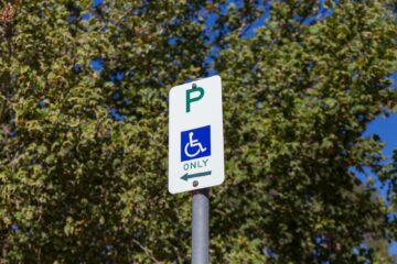 A parking sign indicating accessible parking for people with disabilities, with a wheelchair symbol and an arrow pointing left. Green trees and blue sky are visible in the background.
