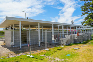 A modern concrete building under construction, surrounded by a temporary metal fence. Stacks of concrete blocks and construction signs are visible, with blue sky and trees in the background.