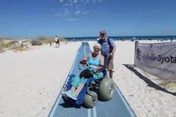 An older woman in a beach wheelchair is being pushed by an older man on a blue mat leading to the ocean. Other people are on the sandy beach; a “Melville Toyota” banner is visible. The sky is mostly clear and sunny.