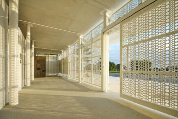 A modern hallway with perforated metal screens casting circular shadows on the floor, white columns, and a view of the outdoors through an open gate.