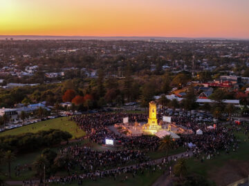A large crowd gathers around a tall war memorial in a park at sunset, with city buildings, trees, and an orange-pink sky visible in the background.