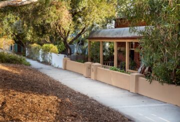 A quiet suburban sidewalk runs alongside a tan fence and a house with a covered porch, surrounded by trees and greenery in soft sunlight.