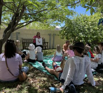 A group of adults and children sit on a green mat under leafy trees, listening to a woman who is reading a book and showing pictures. Sunlight filters through the branches, casting dappled light over the relaxed outdoor gathering.