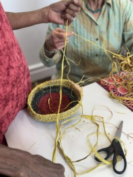 Two people weave a colorful, round basket with yellow fibers at a table. One person holds strands to braid, while the other guides the process. Scissors and a vibrant fabric rest nearby. Only their hands and arms are visible.