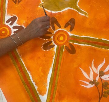 A hand with silver bangles is painting an orange, yellow, and brown abstract artwork with concentric circles, dotted patterns, and floral shapes on a bright orange background.