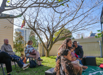A group of people sit outside on chairs and the grass under a tree in a yard, listening attentively. The scene is sunny, with a yellow house, a fence, and a flag visible in the background.