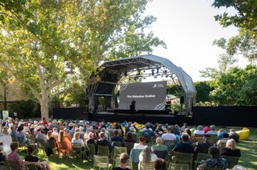 A large audience sits outdoors on chairs facing a stage with a curved canopy. A person speaks at a podium, and a large screen behind them displays “The Walyalup Oration.” The area is shaded by trees and greenery.