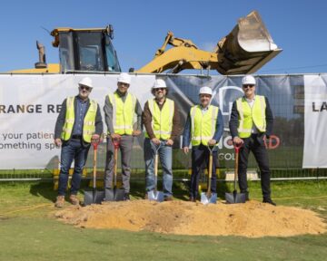 Five men in safety vests and hard hats stand in front of a bulldozer, each holding a shovel at a groundbreaking ceremony on a sunny day. A banner is visible in the background behind them.