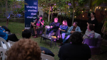 A panel of five people sits outdoors under string lights, engaged in discussion. An audience watches, and a large sign reads, “Sharing stories that change.” Trees and greenery decorate the setting.