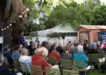 A group of people sit on green chairs outdoors, listening to a speaker in front of a rustic shed. String lights hang from a tree, and leafy trees surround the gathering.