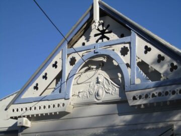 A close-up of a decorative wooden gable on a house, featuring ornate trim, cut-out patterns, and a carved floral and face design, all painted white against a clear blue sky.