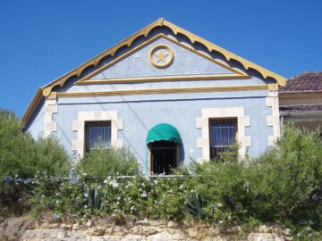 A light blue house with a triangular roof, yellow trim, and a prominent Star of David above the entrance. The house has barred windows, a green awning, and is surrounded by green shrubs and white flowers under a clear blue sky.