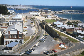 A coastal town with a marina, historic buildings, a curved railway line, parked cars, and a large Australian flag on a flagpole near the water. The scene overlooks the ocean and piers extending into the harbor.