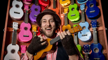 A smiling person wearing a hat and fingerless gloves plays a ukulele in front of a wall filled with colorful ukuleles.