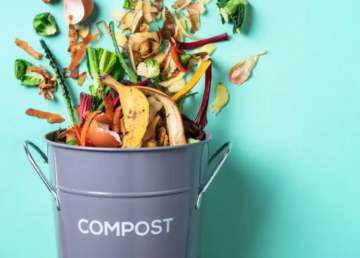 A gray compost bin labeled COMPOST is filled with fruit and vegetable scraps, with some food waste flying above the bin, against a light blue background.