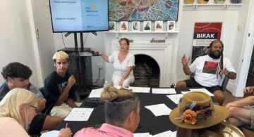 A group of people sits around a table covered with papers, participating in a workshop. A woman points to a screen displaying words, while a man gestures animatedly. Colorful art and photos decorate the room.