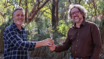 Two smiling men standing outdoors in a wooded area, holding a small plant together. One wears a plaid shirt and the other a brown shirt with glasses. Bright sunlight filters through the trees in the background.
