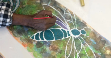 A person with medium brown skin and a plaid shirt paints a blue and white insect with large wings on a textured, multicolored background.