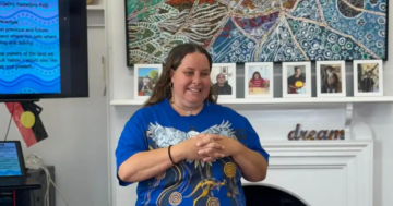 A woman wearing a blue shirt with an artistic design stands smiling in front of a white fireplace, with family photos, Indigenous decorations, and a dream sign on the mantelpiece. A screen with text is visible on the left.