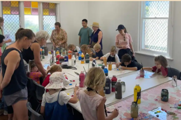 Children and adults gather around tables covered with art supplies, paint bottles, and paper, engaging in a group art activity in a bright, sunlit room with large windows.
