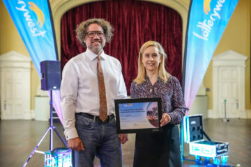 Two people stand indoors smiling at the camera. The woman on the right holds a certificate. Colorful Lotterywest banners are displayed behind them, and the background features a stage with a red curtain.