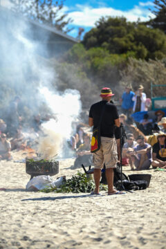 A person stands on a sandy beach speaking into a microphone, facing a group of seated people. Smoke rises from a container with leaves burning. Trees and blue sky are in the background.