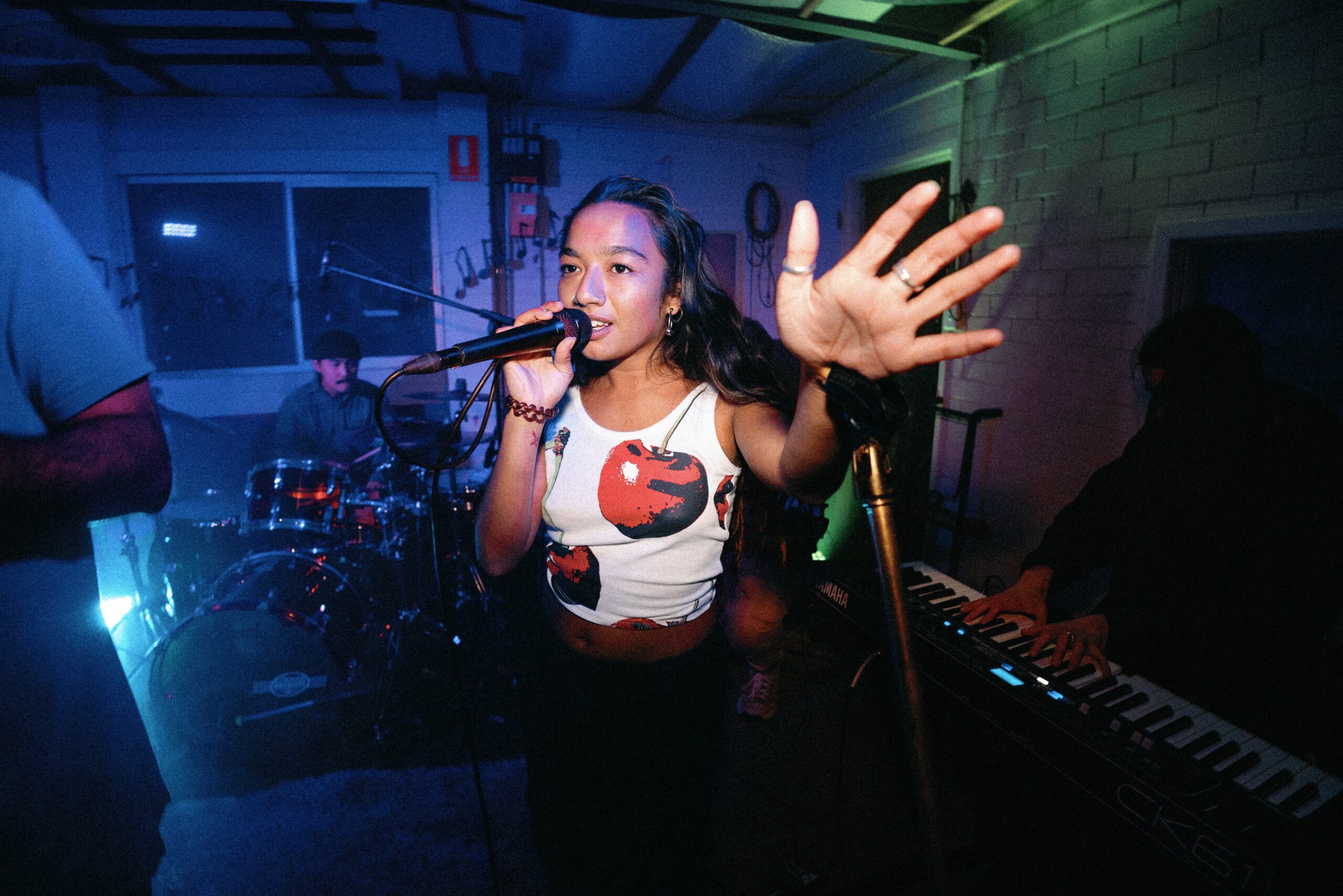 A woman sings into a microphone with her hand raised, performing with a band. A drummer and keyboard player are visible in the background under colorful stage lights.