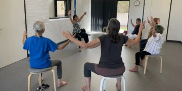 A group of older adults sits on stools in a circle, raising their arms while participating in a seated exercise class in a bright indoor room.