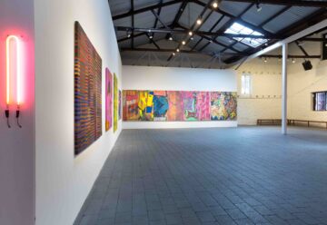A spacious art gallery with high ceilings, featuring colorful abstract paintings on white walls, a neon light fixture to the left, and large windows letting in natural light.