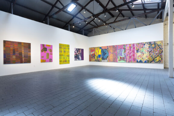 A spacious art gallery with high ceilings displays vibrant abstract paintings on white walls. The floor is gray tile, and natural light shines through windows above, illuminating the colorful artwork.