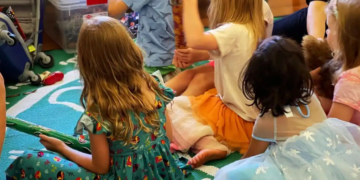 A group of young children sit together on a patterned rug, some holding wrapped gifts. They wear colorful dresses and appear to be engaged in a group activity or celebration.