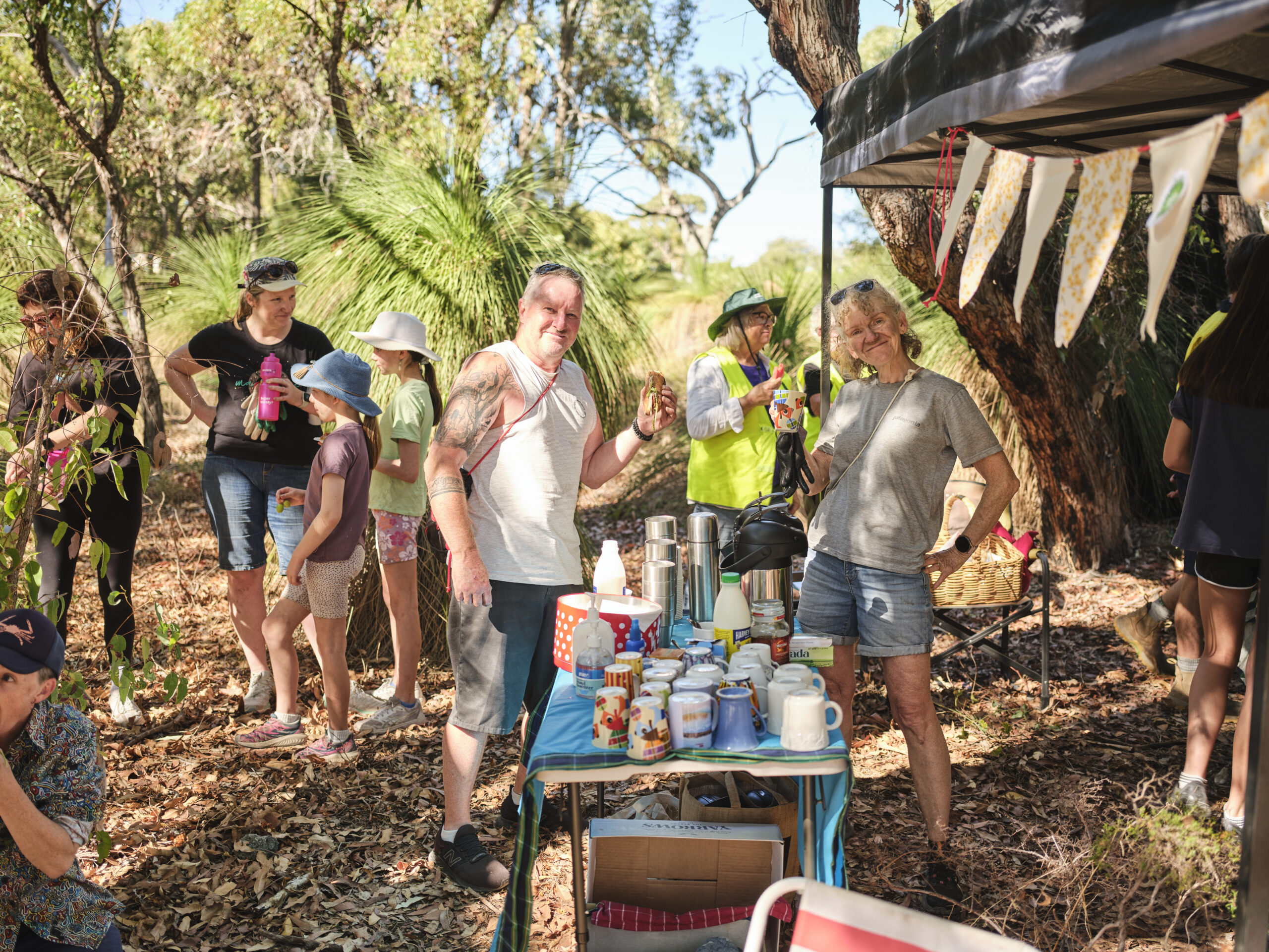 A group of people stand in a sunny, wooded area beside a table with mugs and drinks. Some are smiling at the camera, while others chat or drink. There is a tent with bunting and scattered leaves on the ground.