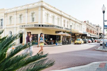 Two women in stylish outfits cross a sunny street lined with parked cars, palm leaves in the foreground, and a cream-colored historic building with Bar & Kitchen signs in the background.