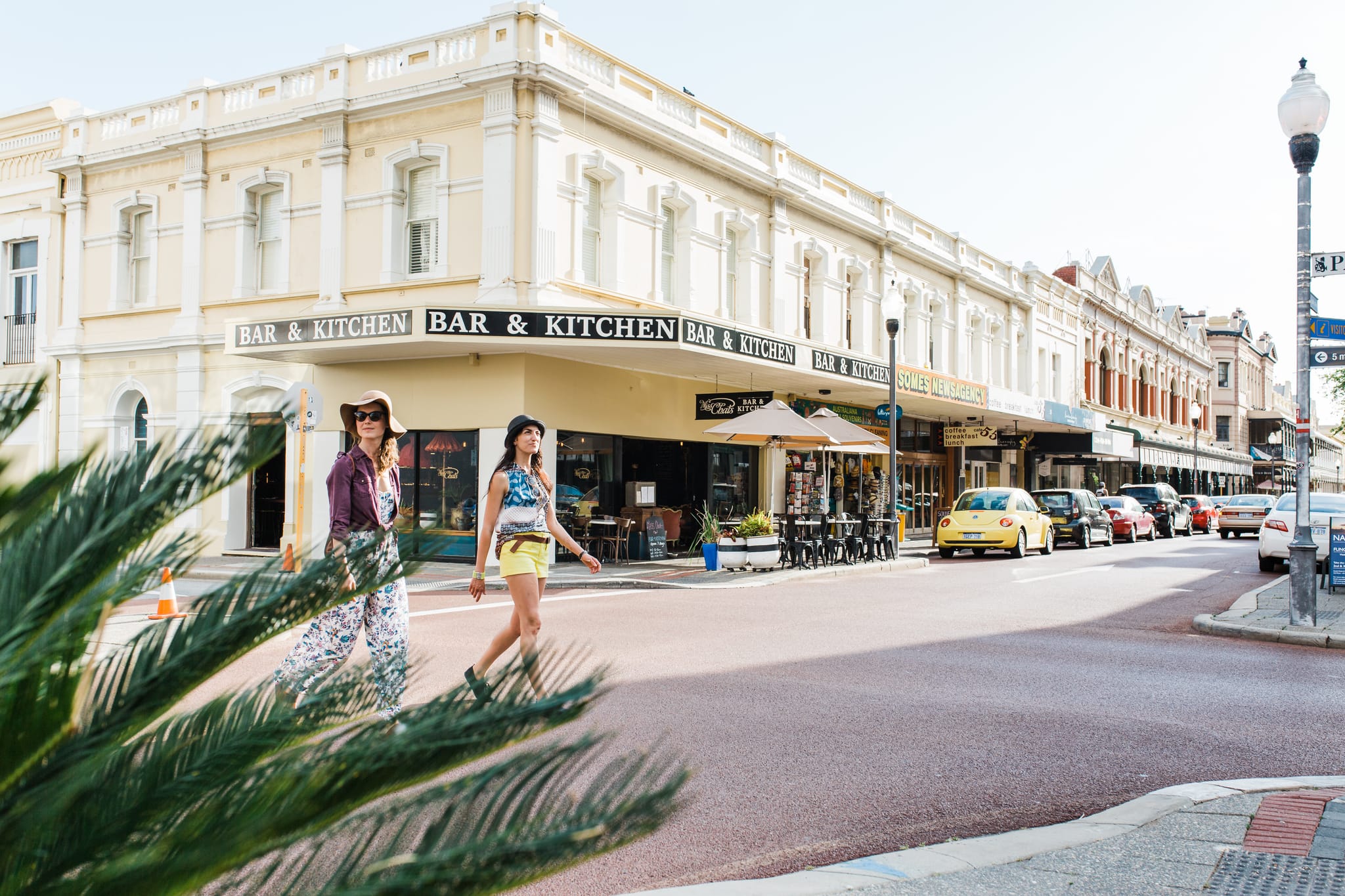 Two women in stylish outfits cross a sunny street lined with parked cars, palm leaves in the foreground, and a cream-colored historic building with Bar & Kitchen signs in the background.