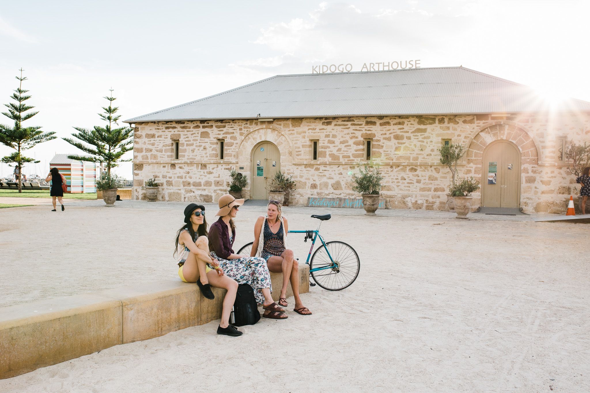 Three women sit and chat on a low wall outside the stone Kidogo Arthouse building, with a bicycle nearby and the sun shining in the background.