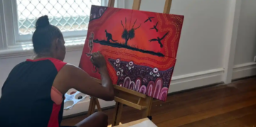 A person with dark hair in a bun paints an Aboriginal-style artwork featuring animals and birds on a canvas set on an easel, inside a sunlit room with wooden floors and a window.
