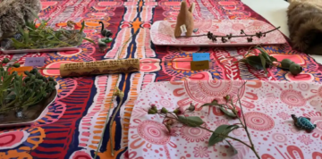 A colorful tablecloth with Aboriginal-inspired patterns holds trays displaying native plants, leaves, twigs, and small labeled objects, including a piece of wood with writing and animal fur at the edges.