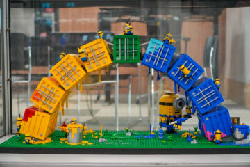 A LEGO scene features Minions building two colorful arches made of yellow and blue crates, with several Minion figures climbing and working on the structures, set on a green base with various construction props.