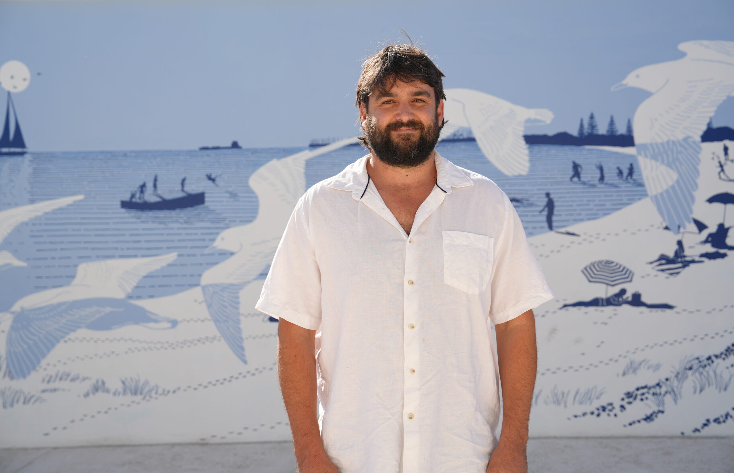 A man with dark hair and a beard, wearing a white shirt, stands in front of a wall mural depicting seagulls, people on the beach, and a boat on the ocean. The scene is drawn in shades of blue and white.