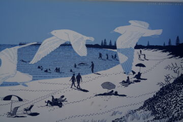 Illustration of a beach scene with people relaxing, swimming, and walking. Large seagulls fly overhead, and beach umbrellas, towels, and footprints are visible on the sand; the artwork uses blue and white tones.