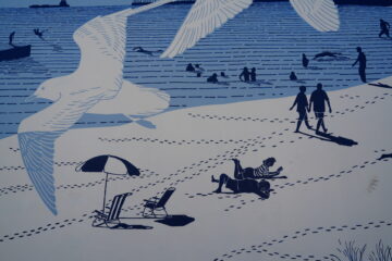 Illustration of a beach scene with people swimming, walking, and relaxing on the sand. Large seagulls fly overhead. An umbrella and beach chairs are set up on the sand, with footprints visible throughout.