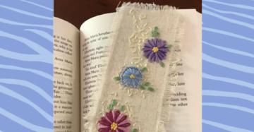 A fabric bookmark with embroidered purple, blue, and pink flowers rests on the pages of an open book. The bookmark has frayed edges and delicate green leaves stitched around the flowers.
