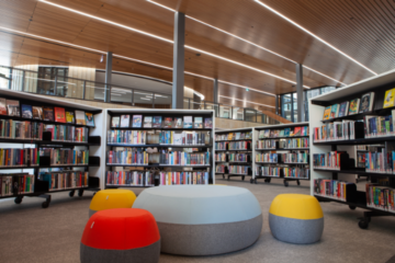 A modern library interior with shelves of books, round cushioned seating in gray, red, and yellow, and a high wooden ceiling with linear lighting. Large windows provide natural light.