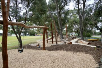 A nature playground with wooden climbing structures, swings, balance beams, and log seating set among trees on woodchip and mulch ground, with grassy fields and more play equipment in the background.