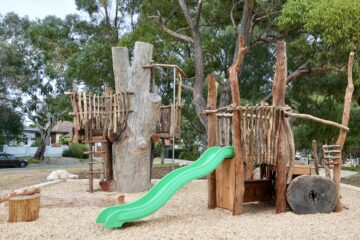 A natural playground with wooden structures, including tree trunks, rope bridges, and a bright green slide, surrounded by trees and wood chips on the ground.