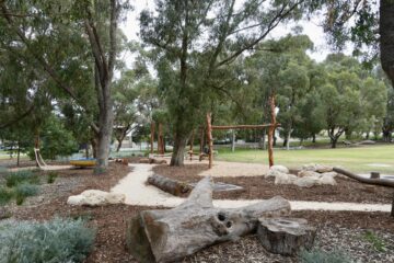 A natural playground with wooden climbing structures, logs, rocks, and trees surrounding a dirt path on a grassy area in a park.