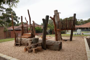 A natural playground featuring large wooden logs, stumps, a wooden platform, and a rope ladder, surrounded by wood chips. Houses with red tile roofs and trees are visible in the background.
