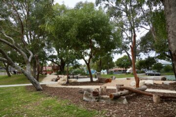A park with large trees, a walking path, picnic tables, logs and rocks arranged as play structures, and houses visible in the background on a cloudy day.