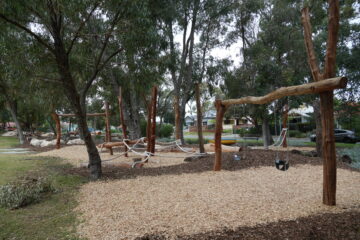 A natural playground with wooden climbing structures, swings, ropes, and wood chip ground cover, surrounded by tall trees and greenery in a suburban park setting.